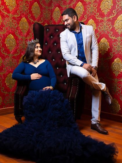 A regal couple's portrait. The mom-to-be is seated on a throne-like chair in a navy blue gown, with her partner looking on affectionately. The rich, patterned wallpaper adds to the opulent feel.