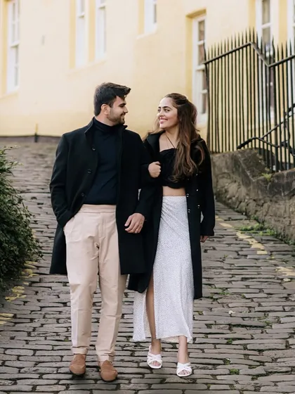 The couple strolls arm-in-arm down a charming cobblestone street in Edinburgh. Their stylish outfits and the historic setting create a classic and sophisticated European romance vibe.