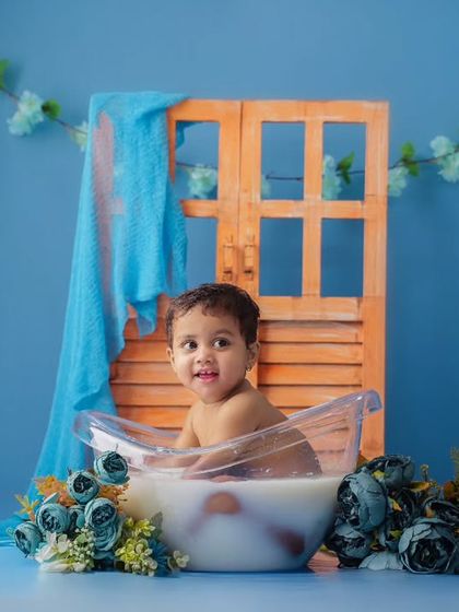 A baby enjoys a milk bath in a clear tub, set against a bright blue background with blue floral decorations.
