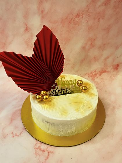 A simple and modern anniversary cake. The design features a textured buttercream finish, a bold red paper fan, and gold spheres for a touch of elegance.