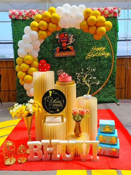A sunny birthday setup with a yellow and white balloon arch against a greenery wall. The plinth arrangement is perfect for the cake and other decorative items.