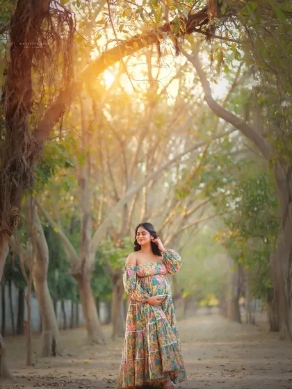 A stunning wide shot in a tree-lined path. The mother-to-be in a floral gown is perfectly framed by nature, with the golden sun creating a heavenly glow.