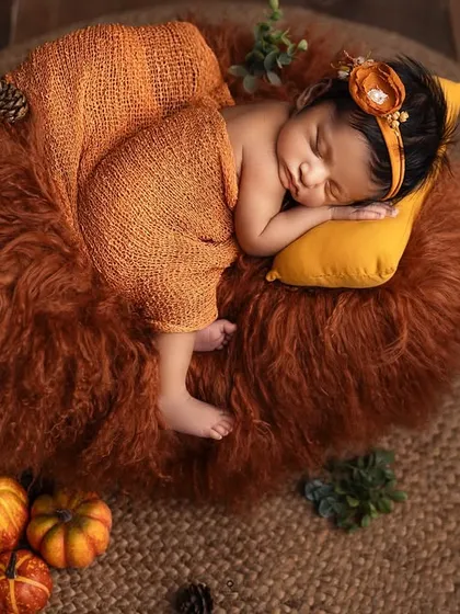 An overhead shot of a newborn nestled in a cozy basket with rich, autumnal colors. The orange wrap, headband, and mini pumpkins create a warm and seasonal portrait.