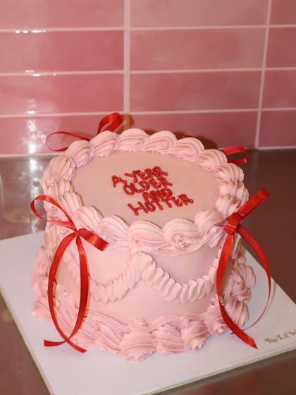 A year older and a year hotter. This pretty pink cake features sassy text, delicate piping, and bright red ribbons for a fun and confident birthday statement.