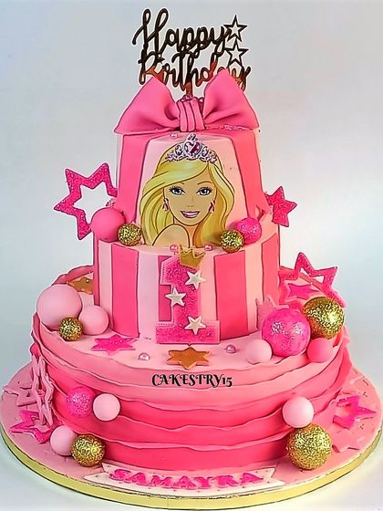 A glamorous three-tier Barbie-themed cake for a first birthday. The pink cake features a Barbie image, sparkling decorative balls, and a large bow topper.