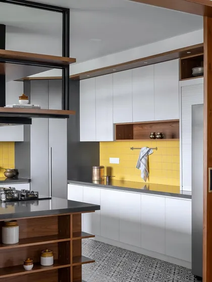 A side view of the 'Vasudhalayah' kitchen island, showcasing the custom open shelving for storage and the sleek black countertop. The suspended chimney unit adds a modern, industrial touch.