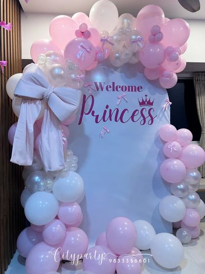 An elegant 'Welcome Princess' decoration for a baby girl's arrival. This home setup features a white arched backdrop, a pink and white balloon arch, and a large fabric bow for a delicate touch.