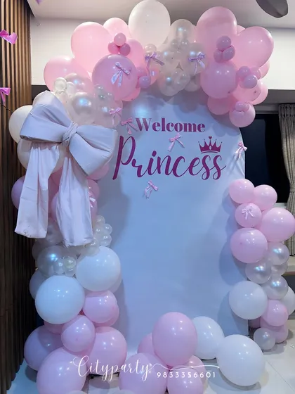 An elegant 'Welcome Princess' decoration for a baby girl's arrival. This home setup features a white arched backdrop, a pink and white balloon arch, and a large fabric bow for a delicate touch.