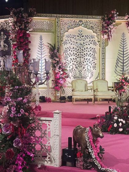 A stage designed with traditional Indian block-print-inspired backdrops and flanked by grand pillars of pink and red flowers. The intricate 'jaali' railing and a Nandi statue add layers of cultural richness to this vibrant and elegant wedding stage.