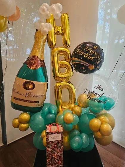 A celebratory balloon bouquet perfect for an adult's birthday. This arrangement features a large champagne bottle foil balloon, 'HBD' letters, and a mix of teal and gold balloons.