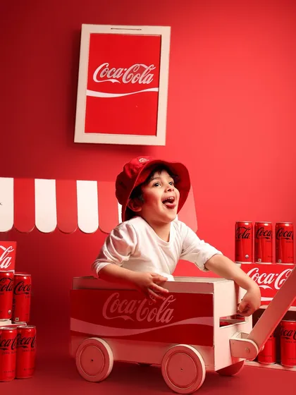 A young boy laughs with joy while sitting inside a Coca-Cola branded cart. The vibrant red and his infectious happiness make this a standout shot from a creative brand-themed session.