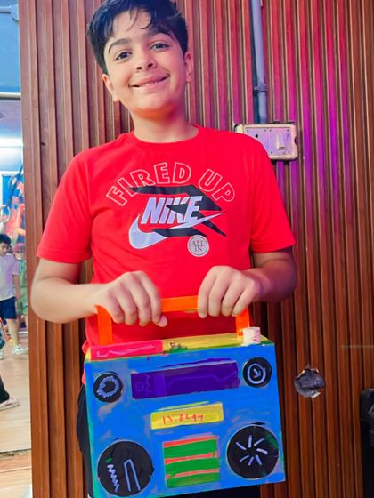A student proudly holds up the colorful boombox he made from cardboard and other craft materials.