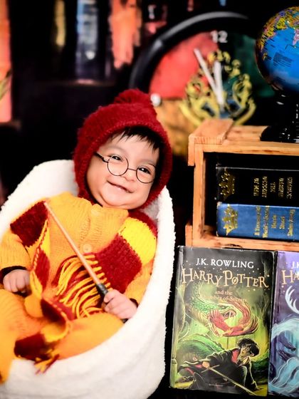 A smiling baby dressed as Harry Potter, holding a wand and sitting next to a stack of the famous books.