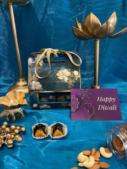 A dark floral carrier box with a window, showcasing the treats inside. This hamper includes dry fruits and traditional Indian sweets like soan papdi.