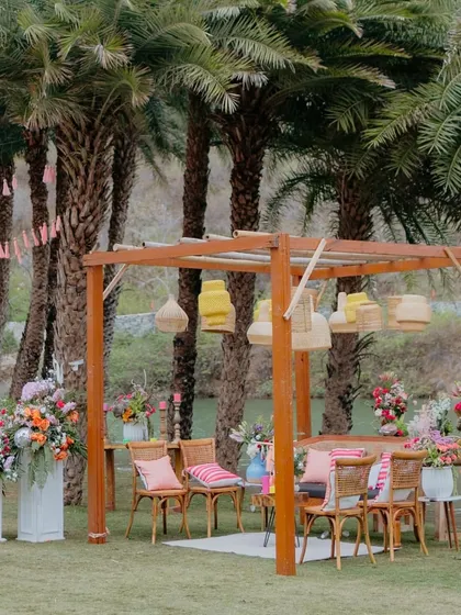 A cozy seating nook at the 'Sunny Sway Soirée'. We used a wooden pergola, hanging cane lamps, and colorful cushions to create a relaxed, boho-chic lounge area by the lake.