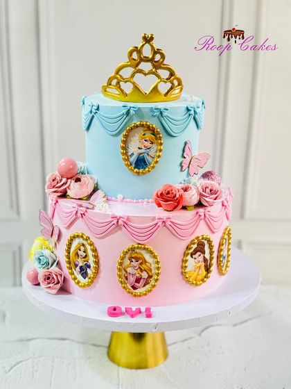 A royal princess cake featuring portraits of Cinderella, Rapunzel, Aurora, and Belle in golden frames. The two-tier pink and blue design is topped with a golden tiara.