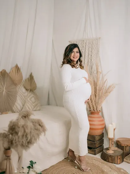 A full-length portrait showcasing the beautiful baby bump. The mother-to-be stands gracefully in a simple white dress, surrounded by a calm, bohemian-style decor.