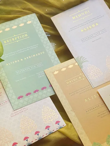 A flatlay of the full invitation suite on pearl-finish paper. The colors range from soft mint green to warm gold and cream, all tied together with delicate lotus and tree illustrations.