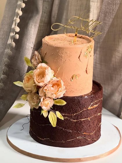 This two-tier anniversary cake combines rustic charm with soft elegance. The rich chocolate bottom tier contrasts beautifully with the peach-toned top, all brought together with delicate florals.