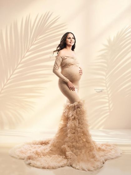 A full-length view of a stunning beige gown with a long, feathery train. The soft, warm lighting and shadow overlays give this studio portrait a gentle and dreamy quality.