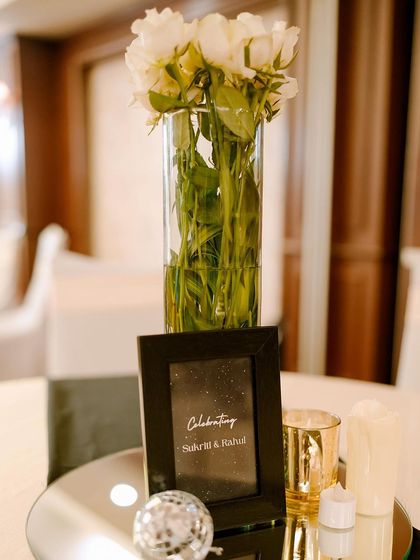 A simple yet elegant centerpiece for a Sangeet dinner table. A small disco ball, a framed "Celebrating" card, and a vase of white roses continue the black, white, and gold theme in a subtle way.