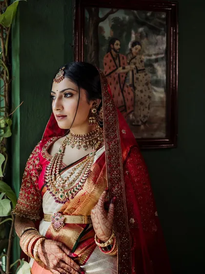 A stunning bridal portrait, with the bride in a red-bordered white saree, her face framed by the window of a suite.