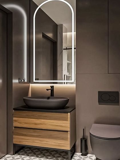 We designed this moody and modern bathroom with dark grey walls and a floating wood vanity. The arched, backlit mirror and black vessel sink add a touch of contemporary elegance to the space.
