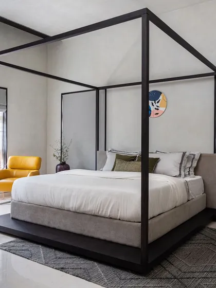 This bedroom features a dramatic four-poster bed frame, creating a modern yet timeless centerpiece. The minimalist design is complemented by a pop of color from the yellow armchair.