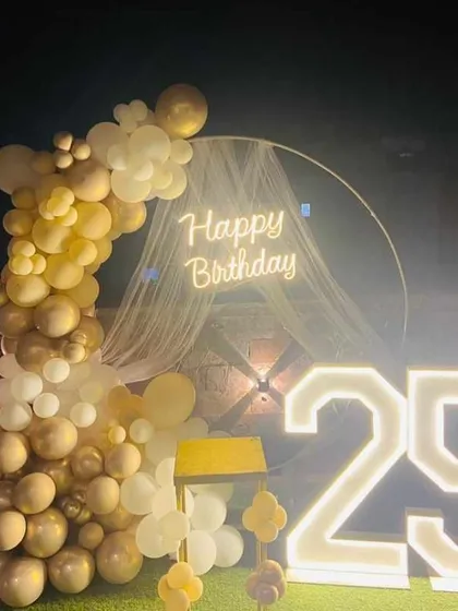 An elegant outdoor setup for a 25th birthday. This decoration includes a half-moon balloon arch in gold and white, a neon sign, and large light-up numbers.