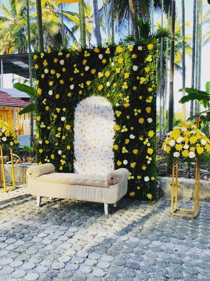 A straight-on shot of the haldi ceremony seating area. The combination of the green floral wall, comfortable bench, and matching floral stands makes for a picturesque and functional setup.