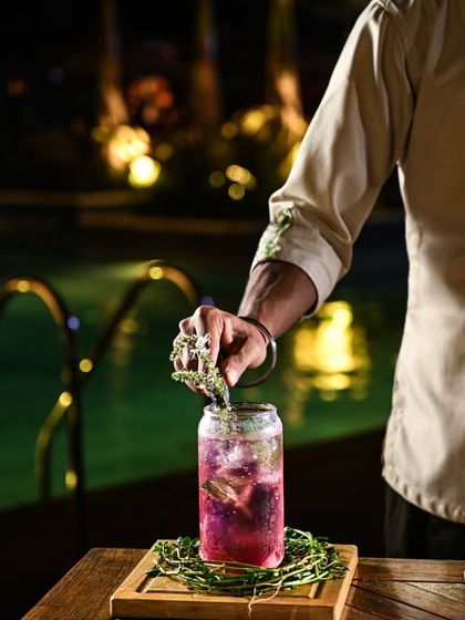 As night falls, our poolside bar Asri comes alive. A bartender carefully garnishes a vibrant, purple-hued cocktail, showcasing the artistry and attention to detail that goes into every drink.