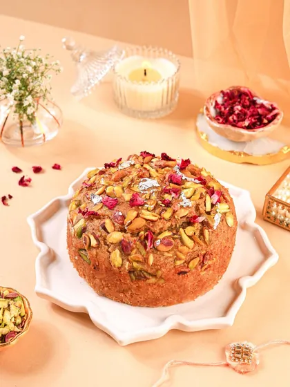 A stunning Mawa cake topped with pistachios, almonds, and rose petals. The soft, peachy background and delicate props create a dreamy and elegant aesthetic.