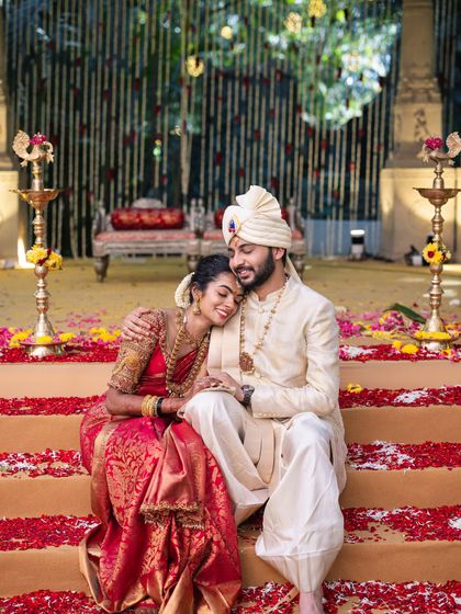 A relaxed moment for a newlywed couple. Seated on steps covered in rose petals, their coordinated red and cream outfits create a romantic and picturesque scene.