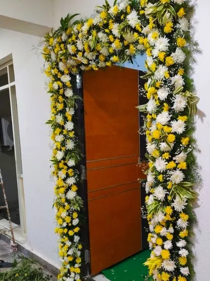 A housewarming flower decoration for a home entrance. The door is framed by an arch of yellow and white flowers with fresh green leaves.
