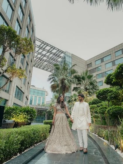 A modern fairytale moment. This wide shot captures the couple walking hand-in-hand through the lush gardens of the JW Marriott Delhi, with the contemporary architecture creating a stunning contrast.