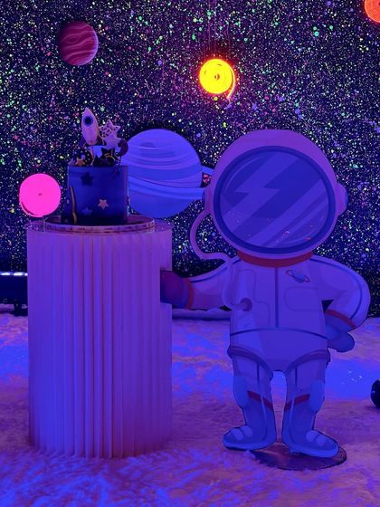 The cake gets a cosmic setting. The space-themed cake is placed on a modern pedestal next to the astronaut prop, in front of the glowing galaxy backdrop.