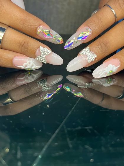 An ethereal and intricate design featuring 3D sculpted flowers, rhinestone bows, and iridescent diamond-shaped gems on long almond nails.