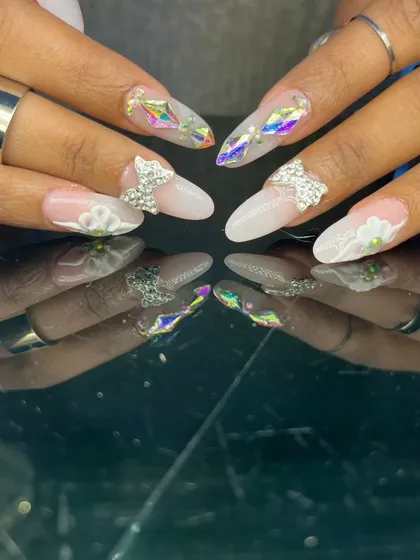 An ethereal and intricate design featuring 3D sculpted flowers, rhinestone bows, and iridescent diamond-shaped gems on long almond nails.