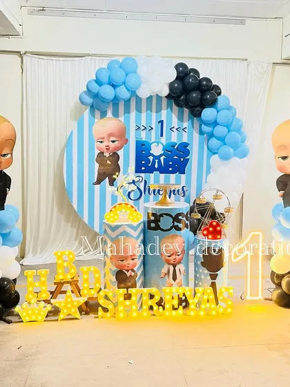 The popular Boss Baby theme for a first birthday party. This decoration includes a striped backdrop, character cutouts, themed pedestals, a blue, black, and white balloon arch, and light-up letters spelling the birthday boy's name.