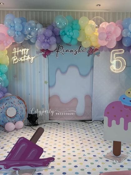 A sweet and colorful candy theme for a 5th birthday party. The setup includes a pastel rainbow balloon arch, a 'Happy Birthday' neon sign, and large props shaped like ice cream, donuts, and marshmallows.