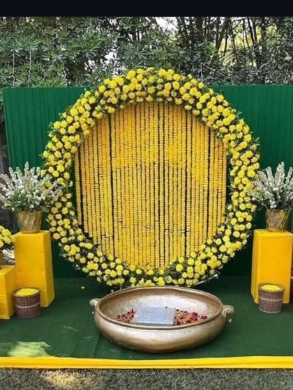 A classic Haldi backdrop for outdoor functions. This setup features a large circular frame filled with strings of yellow marigolds, a green panel background, and a traditional brass urli in the center for the ceremony.