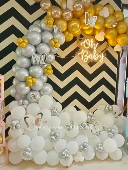 A modern baby shower decoration featuring a half-arch of white, silver, and gold balloons against a black and white chevron backdrop. The setup is accented with butterfly details and 'BABY' blocks.