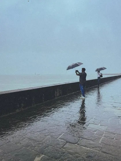 Two people stand against the vast, grey sea at Marine Drive, their umbrellas a shield against the wind and rain. It's a classic Mumbai monsoon frame, full of mood and story.