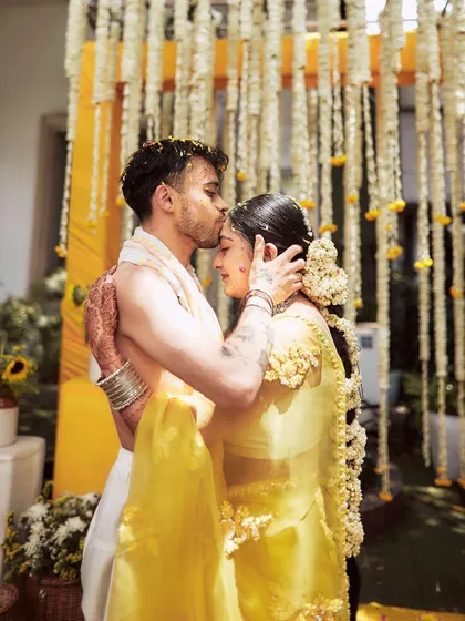 A tender moment where the groom kisses the bride's forehead during their Haldi. The soft yellow tones and floral backdrop add to the romance of the shot.