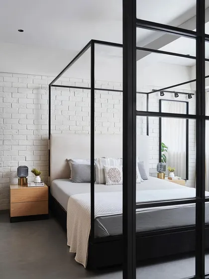Viewing the bedroom through the black-framed glass partition of the study reveals the full composition. The four-poster bed acts as a room-within-a-room, creating a cozy, defined sleeping space that still feels open and light.