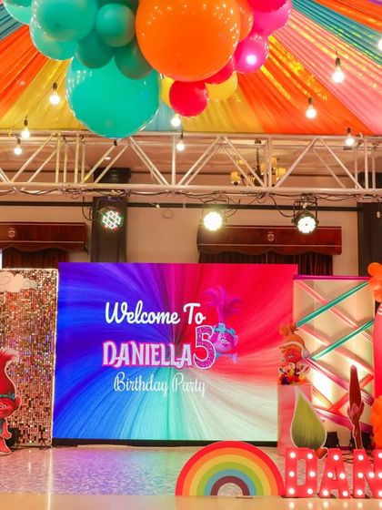 A vibrant and colorful Trolls theme party! The stage is alive with a rainbow-colored ceiling, a bright LED screen backdrop, and character cutouts from the beloved movie.
