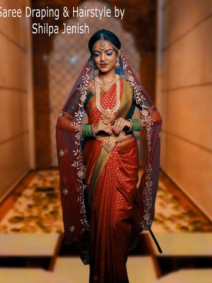 A full-length view showcasing my saree draping and hairstyling services. The bride looks regal in her red silk saree, draped perfectly to highlight her figure, and a traditional floral braid.