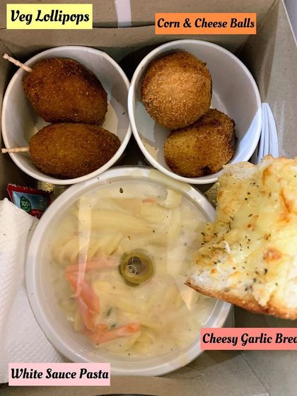 A delicious party meal box featuring white sauce pasta, cheesy garlic bread, veg lollipops, and corn cheese balls. A perfect Italian-themed treat for any celebration.