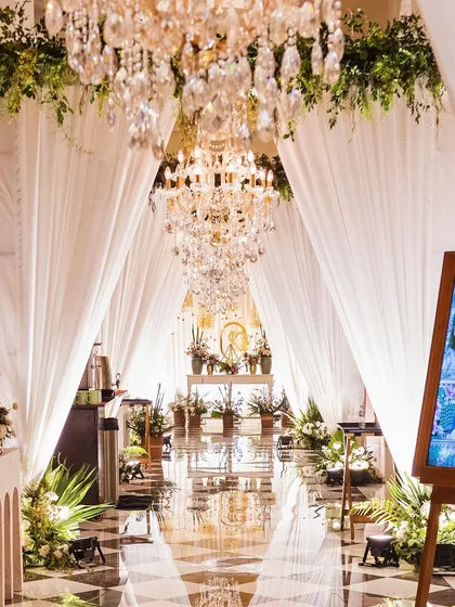 An elegant and grand entrance for an indoor Mehendi function. The design uses white drapes, lush greenery, and magnificent crystal chandeliers to create a sophisticated and dreamy pathway.