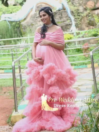 A full-length view of the off-the-shoulder pink ruffled gown in an outdoor setting. The tiered ruffles add beautiful texture and volume, creating an elegant and memorable maternity look.